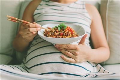 Pregnant woman reclining on a couch with a noodle bowl resting on her belly and chopsticks in her hand