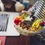 Close-up of person holding up a salad bowl while sitting at desk in front of laptop