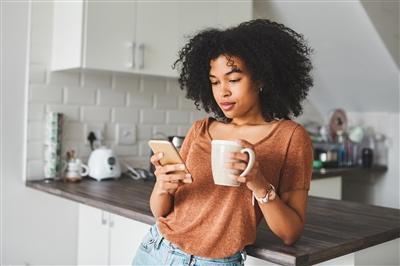 Woman leaning against kitchen counter, holding coffee cup and looking at smart phone