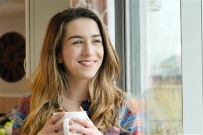 Young woman holding coffee mug looking out living room window with optimism