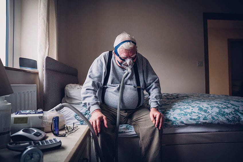 Older adult man seated on bedside with CPAP gear on his head