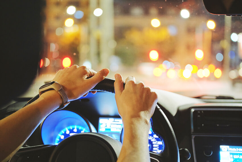 Close up of driver hands holding steering wheel driving car with blurred city street lights on background at night