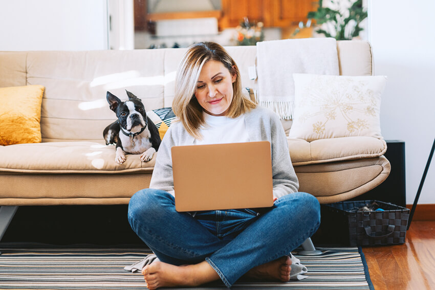 Woman sitting on the floor on her laptop with a dog on the couch behind her. 